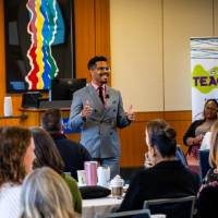 Man in a suit standing in a large conference room, banner that reads "Teach-In" behind him